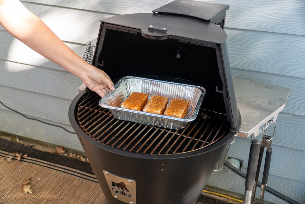 an aluminum tray with three blocks of seasoned cream cheese being placed in the smoker