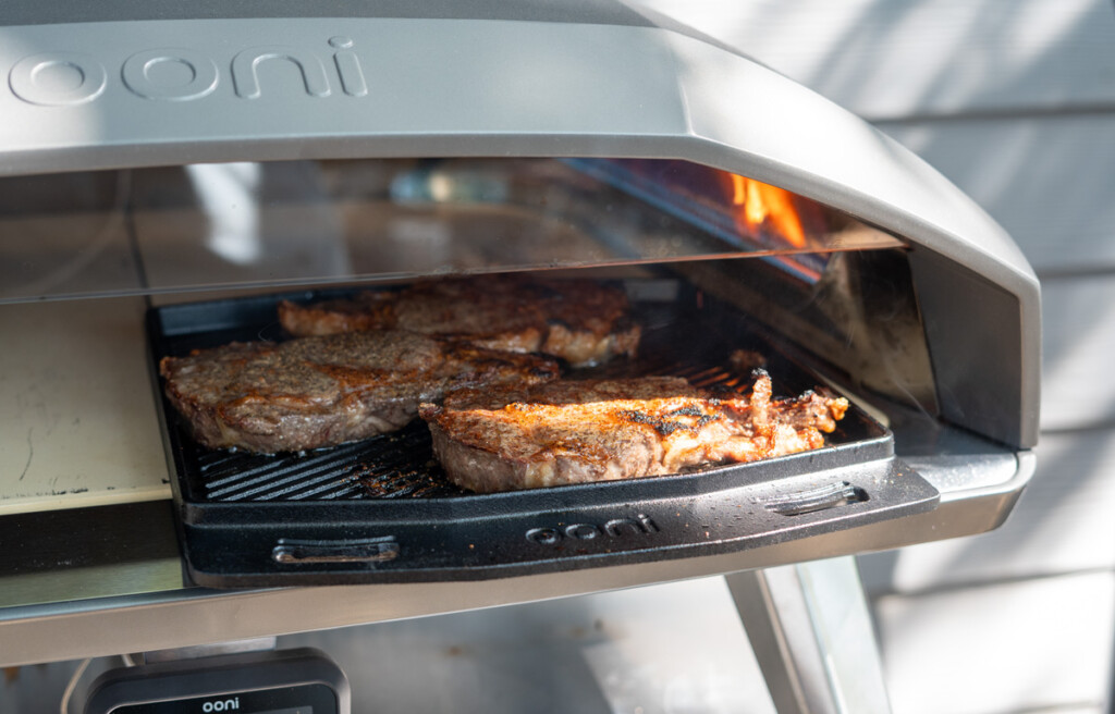 Juicy steaks sizzling on a cast iron griddle inside an Ooni pizza oven, showcasing one of the best things to cook in a pizza oven besides pizza for a perfect seared crust and smoky flavor.
