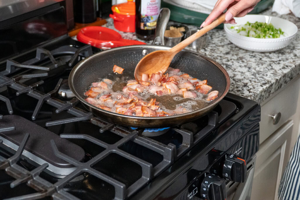 bacon in a frying pan being stirred with a wooden spoon