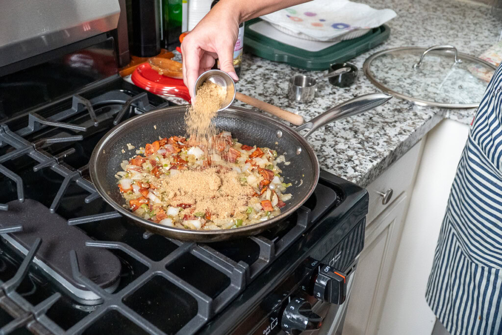a hand tipping a small container of brown sugar into a fry pan