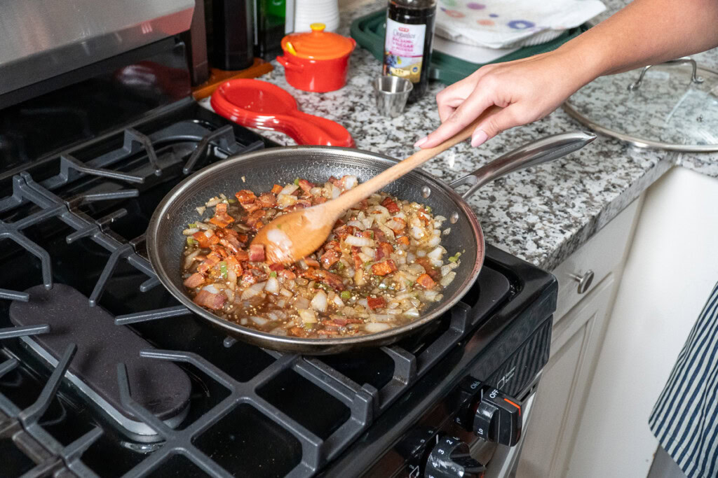 a person stirring a pan of bacon and onions with a wooden spoon