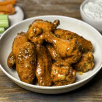 Bowl of smoked buffalo wings coated in glossy orange sauce, served with celery, carrots, and a bowl of ranch dip on a wooden table.