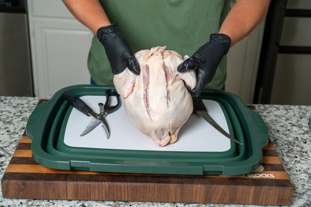 a person hold a whole raw chicken up, with two cuts down either side of the backbone