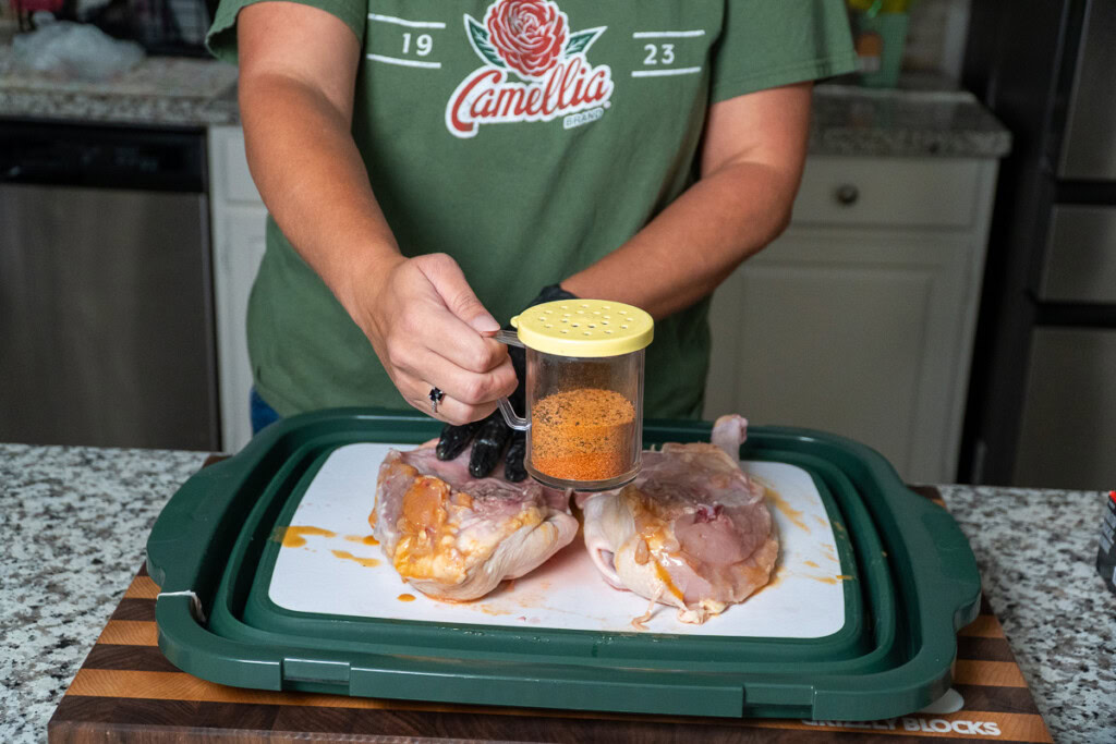 a person with a halved raw chicken and a shaker of seasoning, ready to sprinkle
