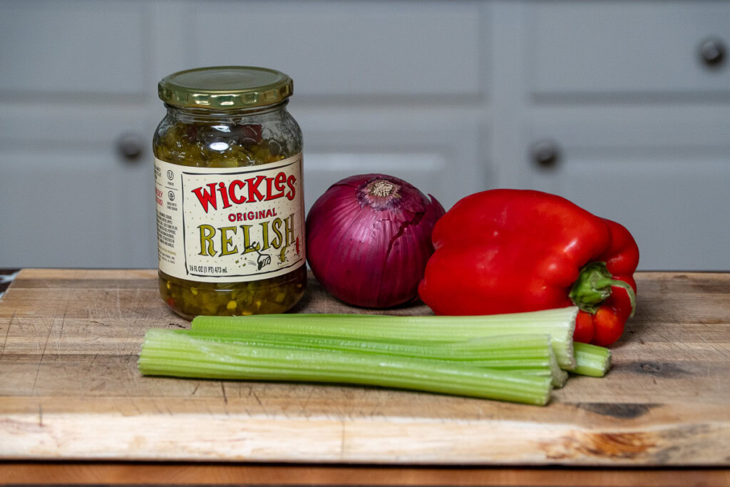 celery, red pepper, red onion and a jar of pickle relish displayed on a wooden board