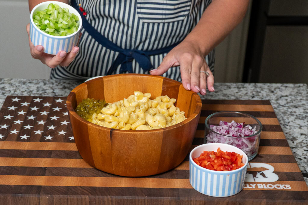 a wooden bowl of cooked pasta, with a bowl of celery being tipped in