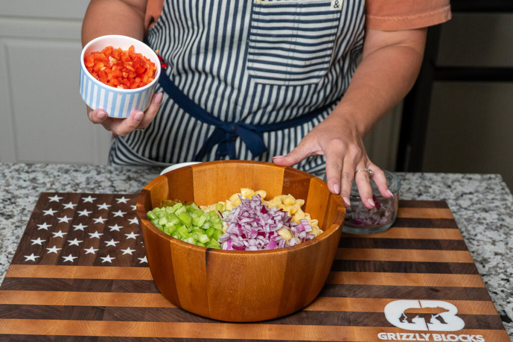 a wooden bowl of cooked pasta with a bowl of diced red pepper being tipped in