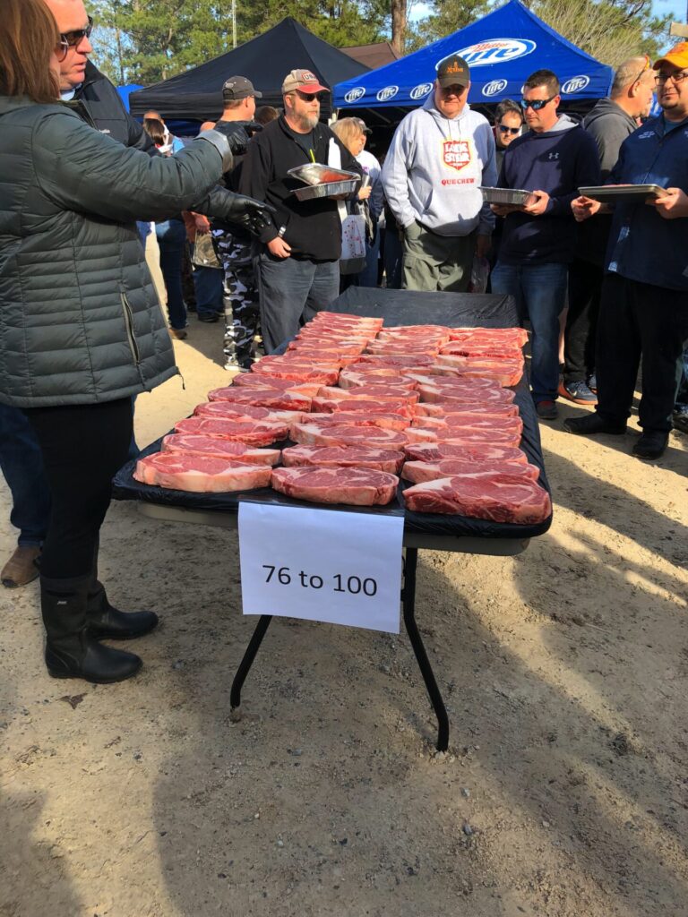 A group of people stand around a table outdoors covered with raw SCA Steak steaks. A sign on the table reads 76 to 100. Some people are holding trays, and tents are visible in the background.