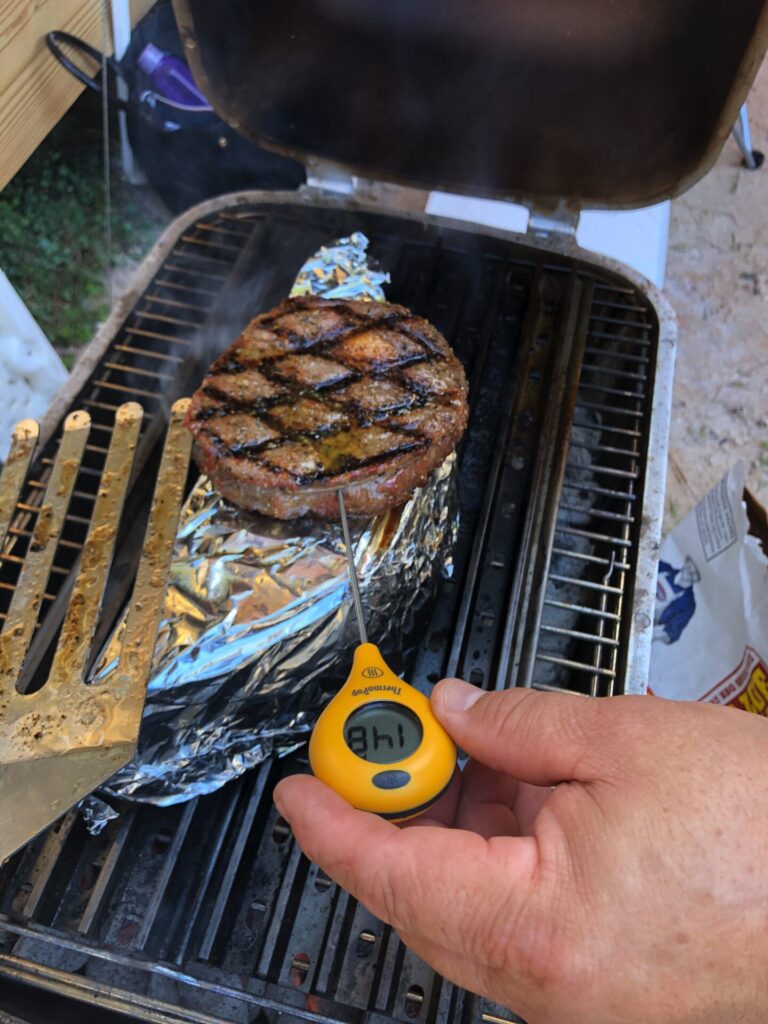 A hand checks the temperature of a grilled hamburger patty with a digital thermometer reading 181°F. The patty sits on a foil-covered grill, SCA Steak style, while a metal spatula rests nearby and smoke rises from the grill.