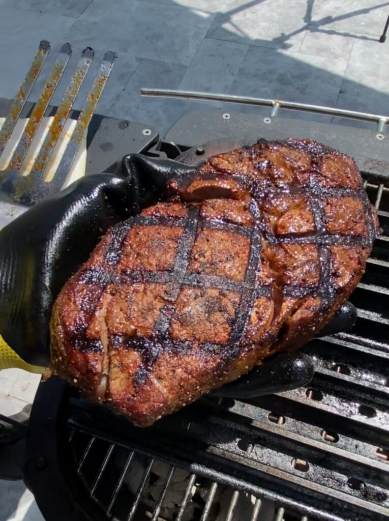 An SCA Steak with prominent crosshatch grill marks is held above a barbecue grill by a person wearing a black heat-resistant glove. A metal spatula rests nearby, hinting at a perfectly executed grilling technique.