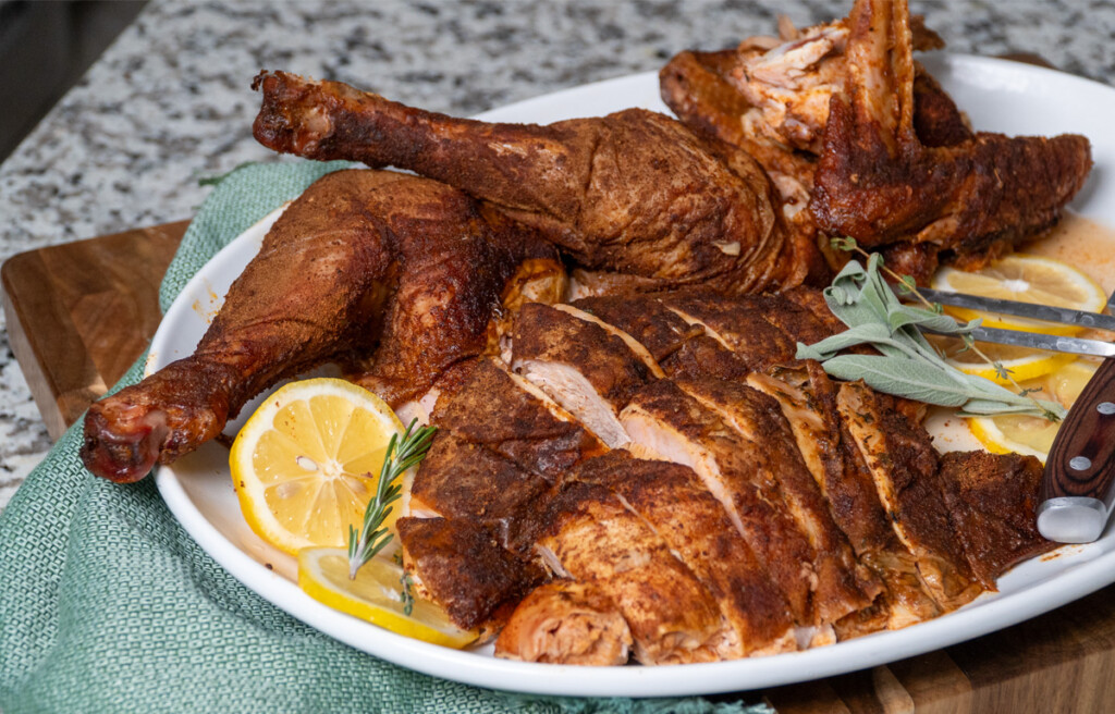 Smoked Cajun turkey carved and served on a white platter with lemon slices, fresh rosemary, and sage garnish, placed on a green napkin on a wooden surface.