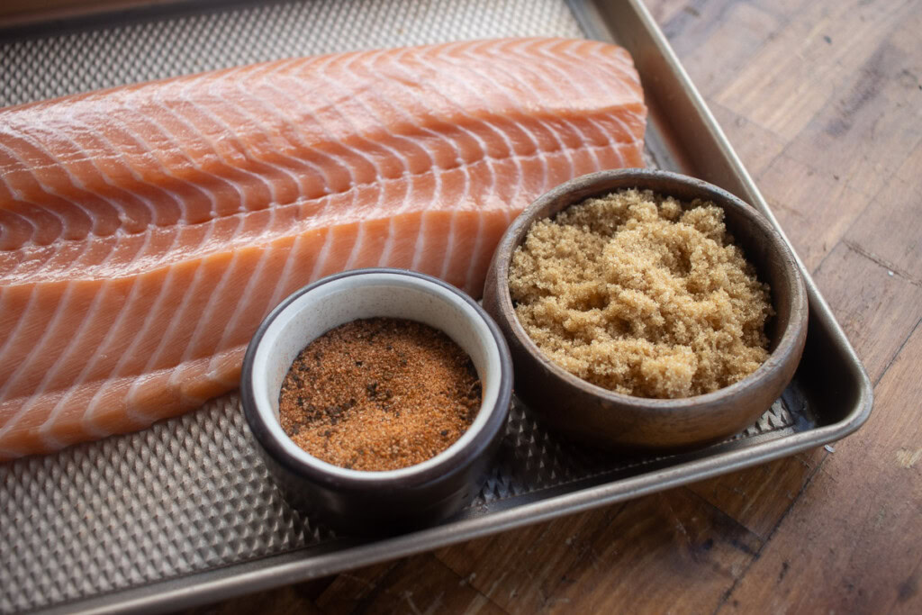 a salmon filet, a bowl of brown sugar and a bowl of bbq rub on a tray