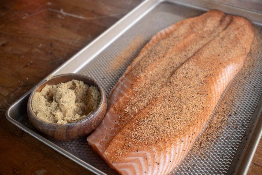 a raw seasoned salmon filet with a bowl of brown sugar next to it