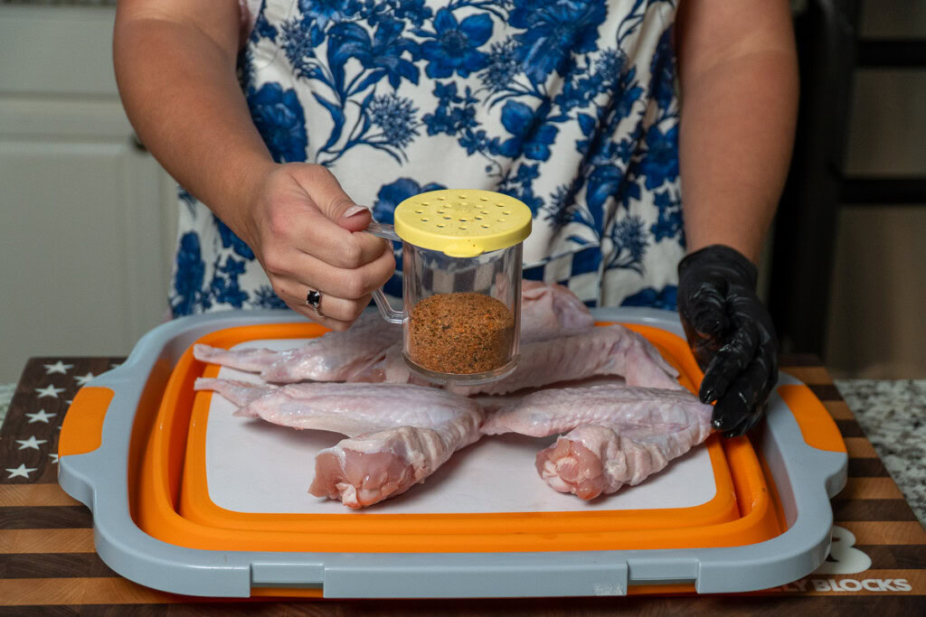a person wearing black gloves holding a shaker of seasong preparing to season raw turkey wings
