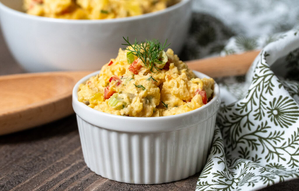 Creamy Southern potato salad served in a white ramekin, garnished with fresh dill and surrounded by a wooden spoon and patterned cloth napkin on a rustic wood surface.
