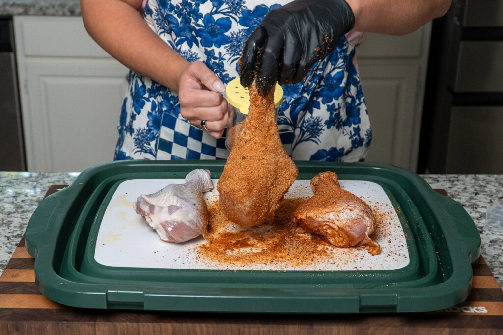 a person with black gloves holding a turkey leg and with a shaker, putting seasoning on it