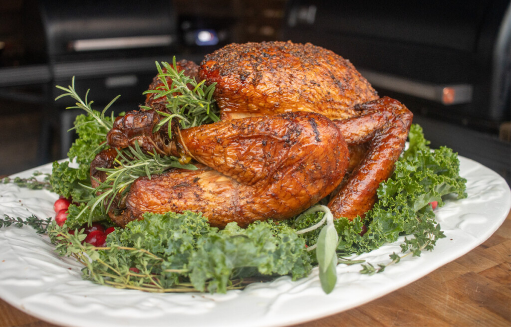 A beautifully smoked turkey on a white platter garnished with fresh herbs such as rosemary, thyme, and kale, with a rich, golden-brown, crispy skin, presented in a rustic setting.
