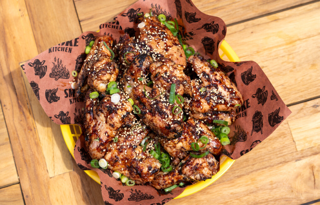 0-400 chicken wings garnished with sesame seeds and sliced green onions, served on brown parchment paper with barbecue-themed graphics, placed in a yellow basket on a wooden table.