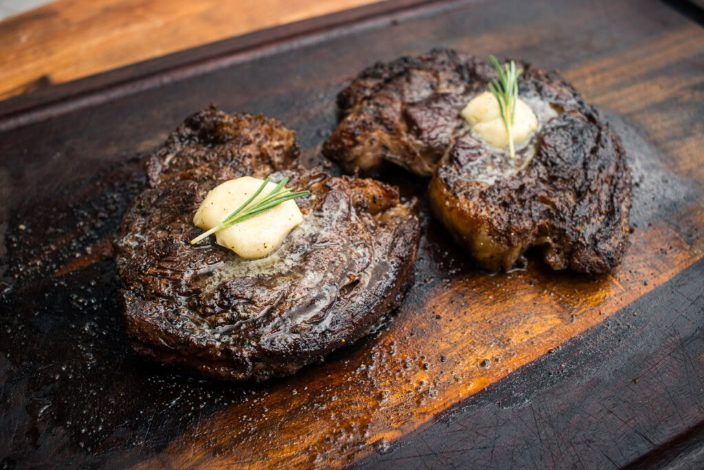 two chuck eye steaks with rounds of butter on top. resting on a wooden board