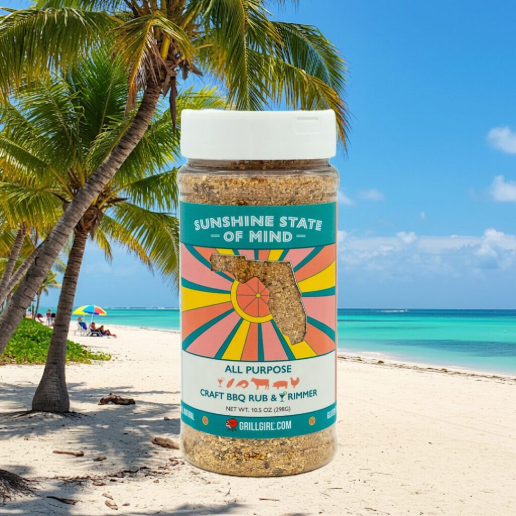 A jar of Sunshine State of Mind all-purpose BBQ rub is shown superimposed on a tropical beach scene, perfect for bringing holiday vibes to your next smoke-fried turkey with palm trees, white sand, blue ocean, and a colorful umbrella in the background.