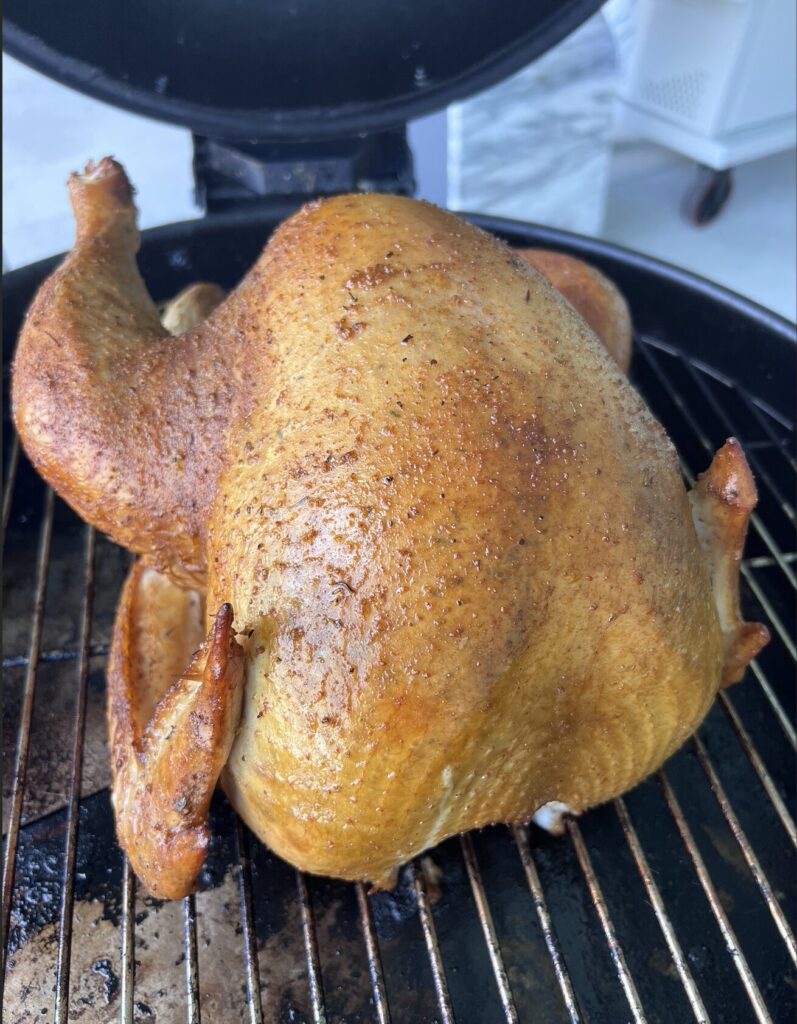A whole, golden-brown chicken is standing upright on a grill inside a barbecue smoker, being cooked outdoors—a true Holiday Obsession, with skin that's crispy, evenly roasted, and perfectly seasoned.