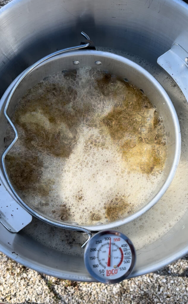 A large metal pot filled with bubbling hot oil is frying food outdoors on a gravel surface. A thermometer tracks the heat as a round basket holds what could be a Holiday Obsession favorite—perhaps even a Smoke-Fried Turkey sizzling to perfection.