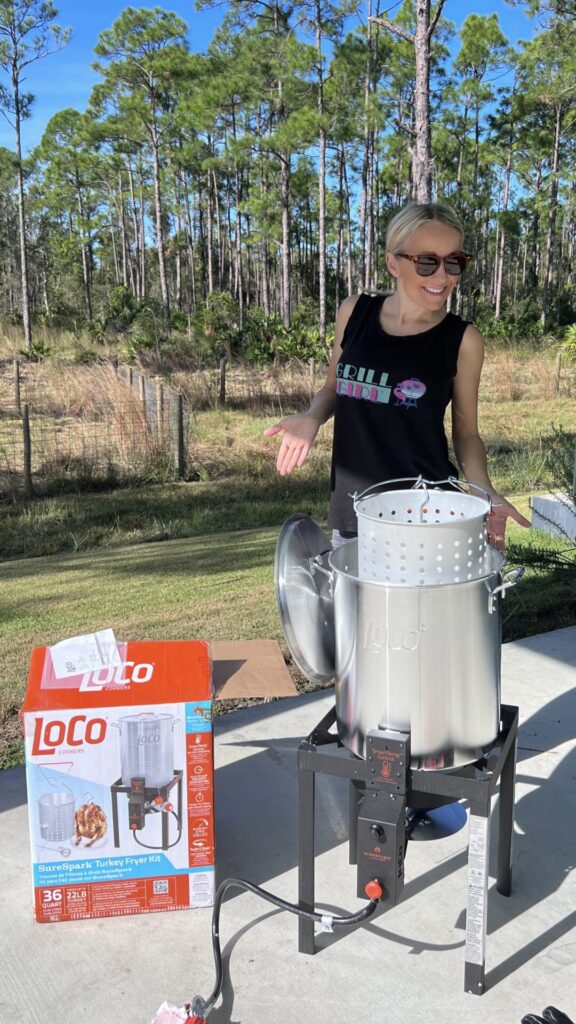 A smiling woman stands outdoors next to a shiny metal turkey fryer and an open LoCo brand turkey fryer kit box, ready for some fried turkey fun, with trees and grass in the background on a sunny day.