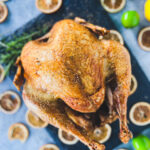 A whole holiday turkey sits on a dark serving board, surrounded by dried lemon slices and green and yellow fruits on a gray surface. The turkey is golden brown and crispy, perfect for any festive gathering.