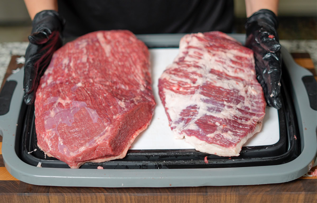 Two separated brisket muscles—the flat on the left and the point on the right—resting on a cutting board. A person wearing black nitrile gloves holds each piece, showcasing the difference in texture and marbling between the lean flat and the fattier point.