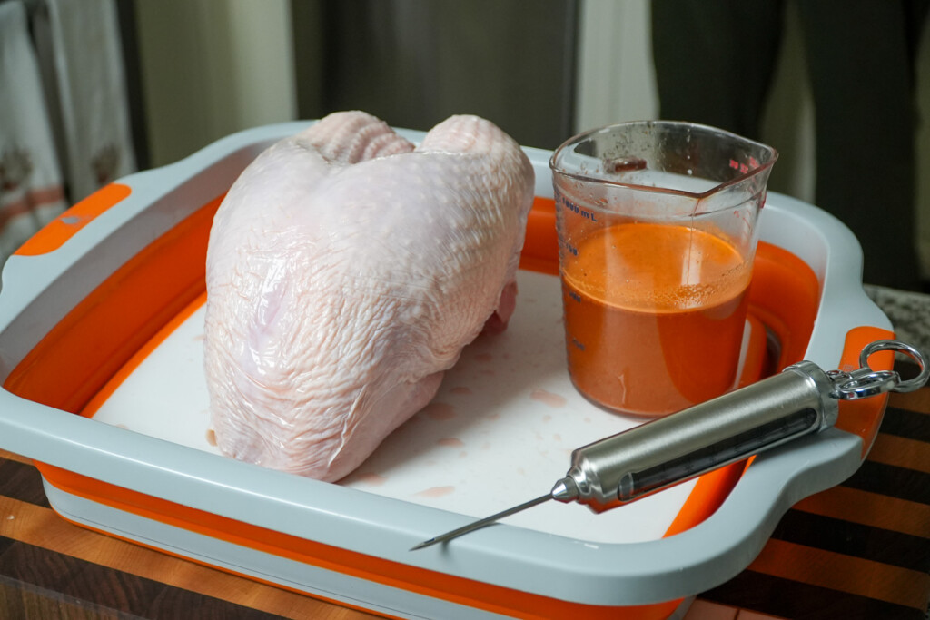a raw turkey breast, a cup of injector and and injector on a grey tray