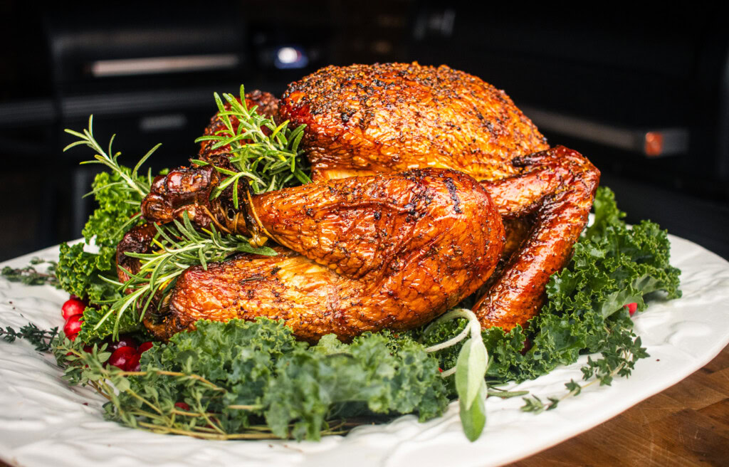Smoked whole turkey on a white platter garnished with fresh rosemary, thyme, and kale, cooked on a pellet grill with golden crispy skin.