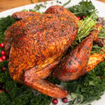 “Whole roasted turkey with deeply browned, crispy skin after a dry brine, served on a platter with rosemary, kale, and cranberries.