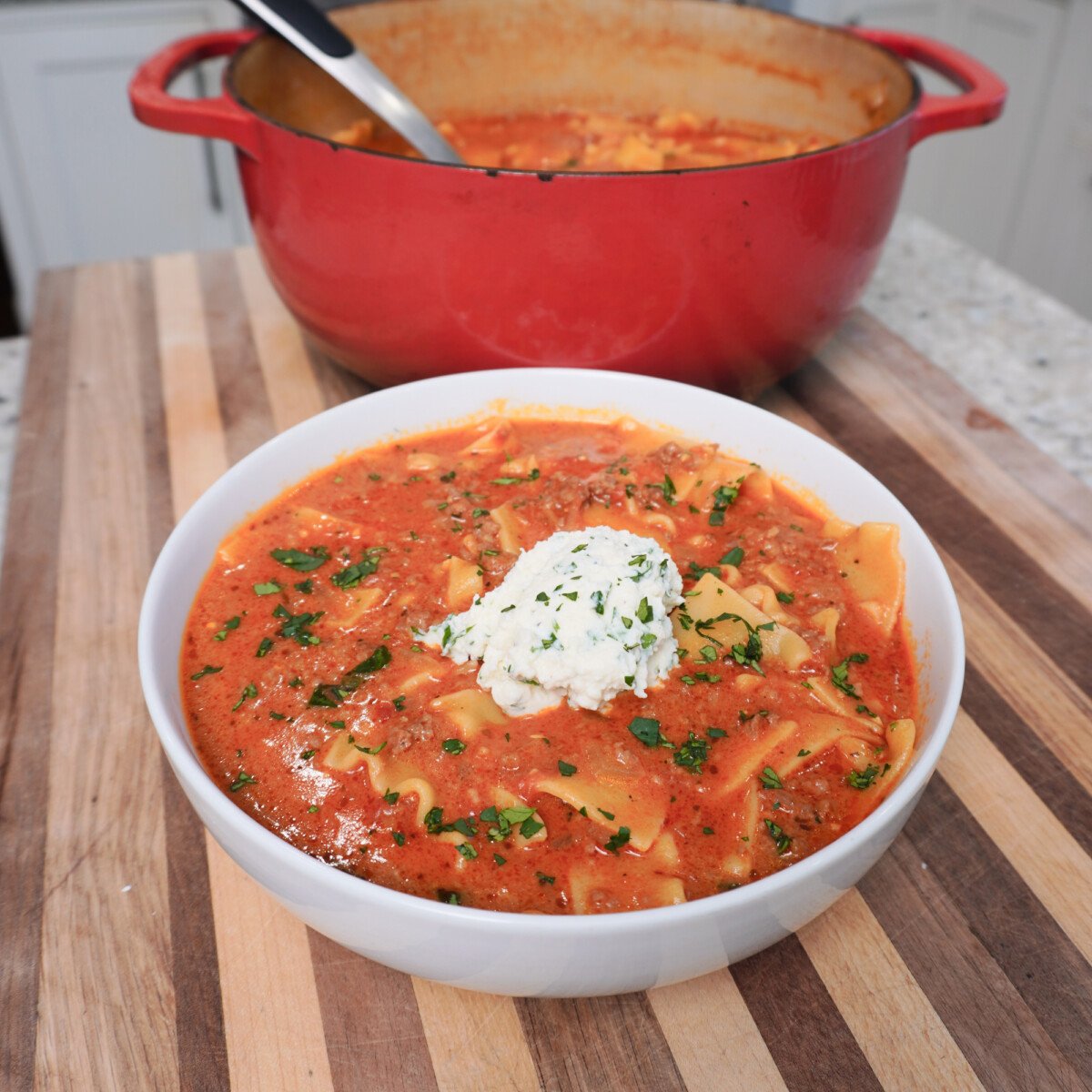 Lasagna soup plated in a white bowl on a stripped cutting board with ricotta cheese on top.