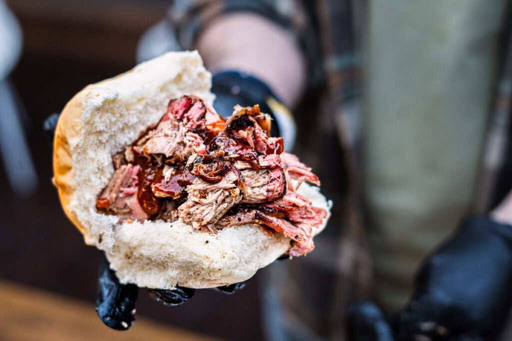 Hand holding a roll filled with pulled smoked lamb shoulder and BBQ sauce.
