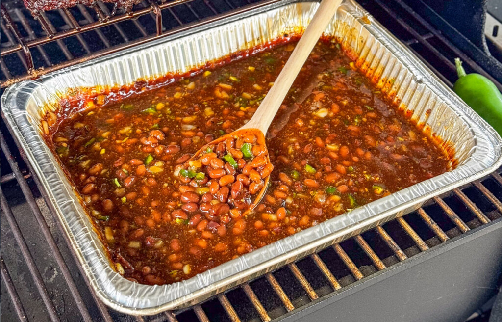 Smoked baked beans cooking in an aluminum pan on a smoker grate, stirred with a wooden spoon. Beans are thick, saucy, and flecked with diced jalapeño and onion.