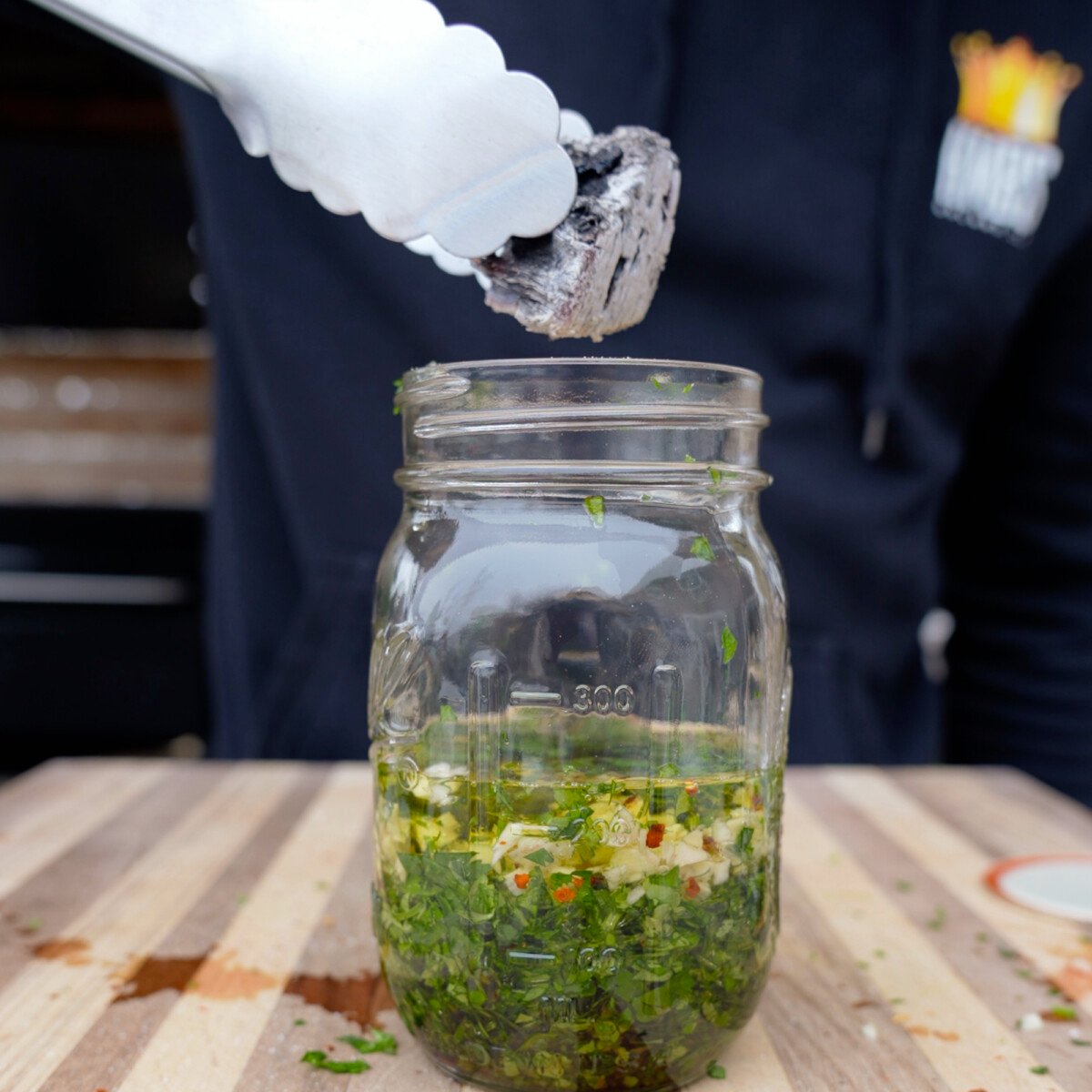 Adding a hot and charred wood chunk to a mason jar full of chimichurri ingrendients for a smoked chimichurri recipe.