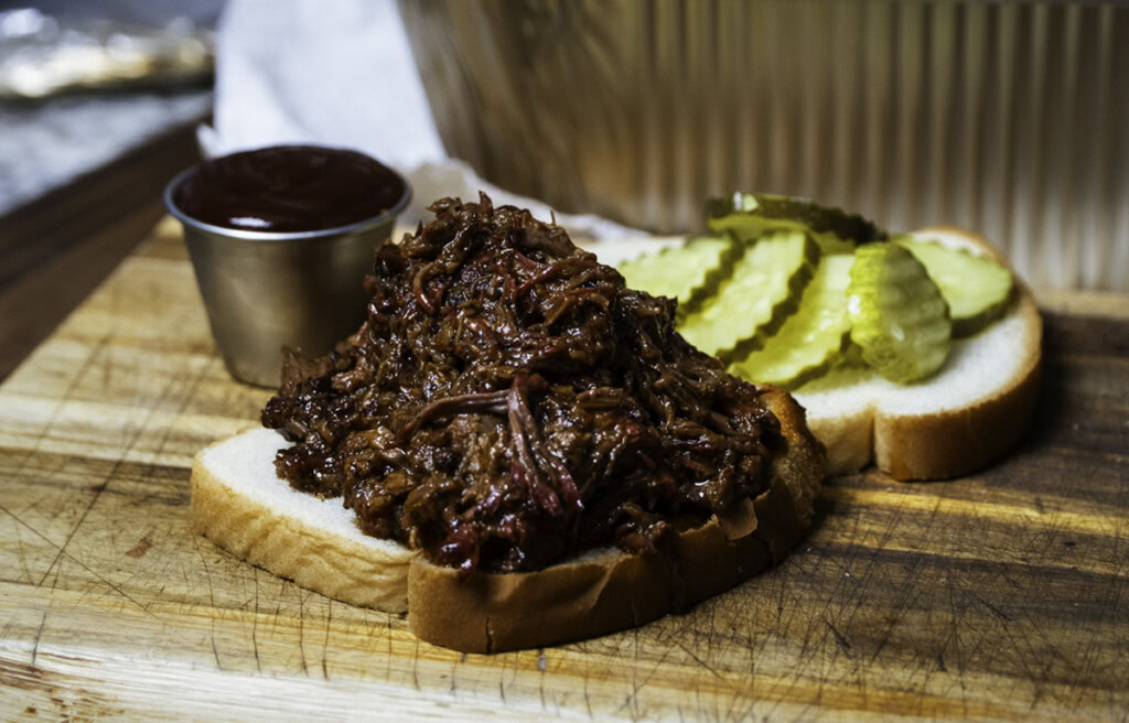 Pulled smoked chuck roast piled onto a slice of white bread, served open-faced with barbecue sauce and pickles on the side.
