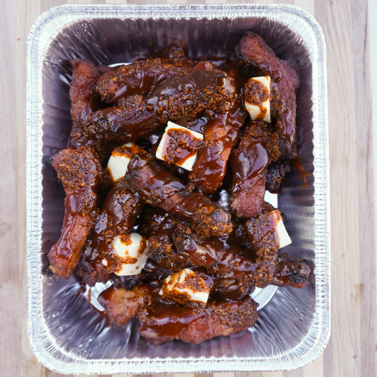 smoked ribs covered in bbq sauce, chili crunch and butter in a foil pan.
