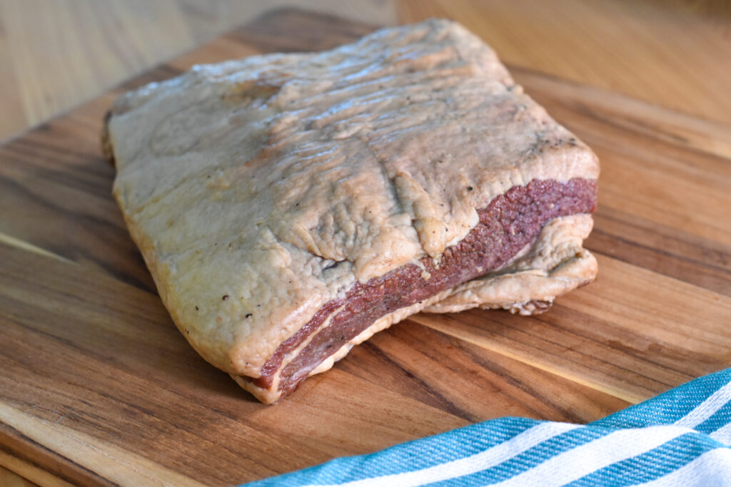 A slab of uncooked brisket flat with a thick layer of fat sits on a wooden cutting board, next to a blue and white striped cloth—perfect for your next beef bacon recipe.