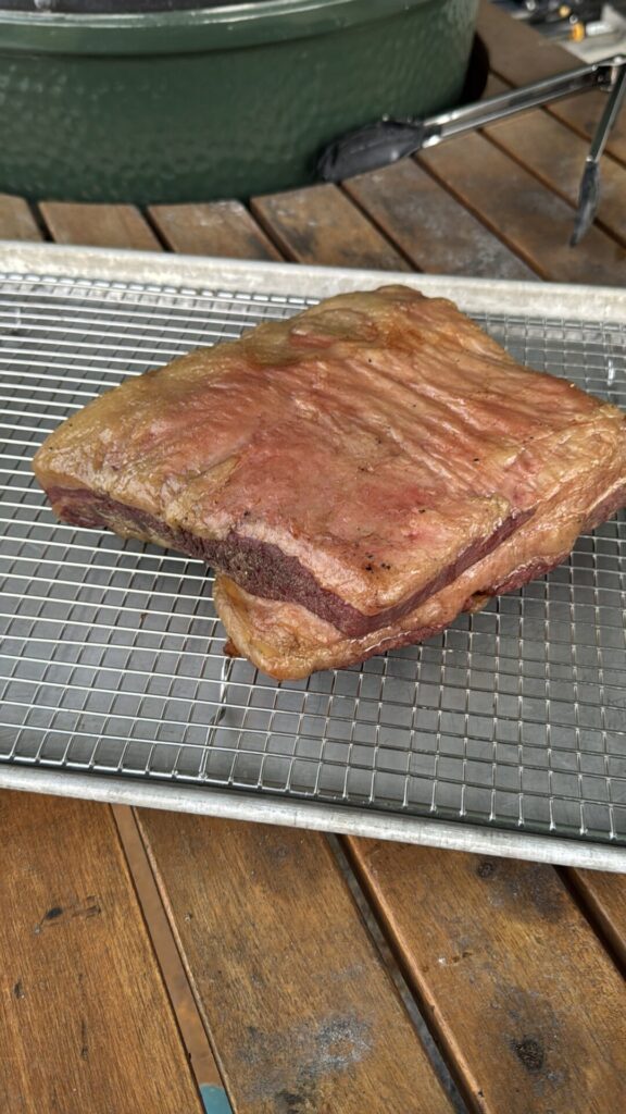 A slab of smoked brisket flat rests on a wire rack over a baking sheet, set on a wooden table. A green grill and metal tongs are visible in the background.