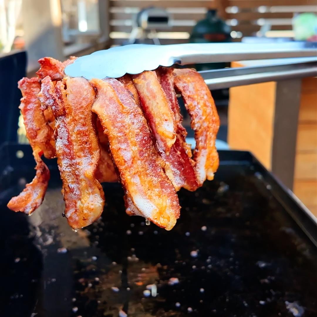 greasy cooked bacon on a griddle held in the air by tongs.