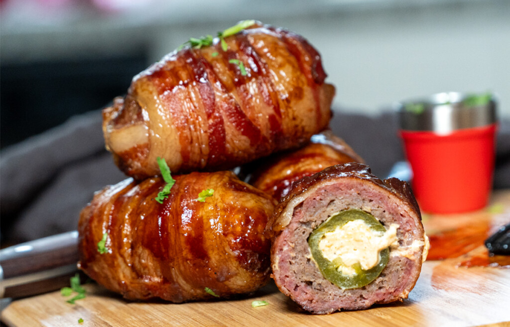 Close-up of smoked Armadillo Eggs—jalapeños stuffed with cheese, wrapped in seasoned sausage, and covered in crispy bacon, glazed with barbecue sauce and garnished with fresh herbs on a wooden cutting board.