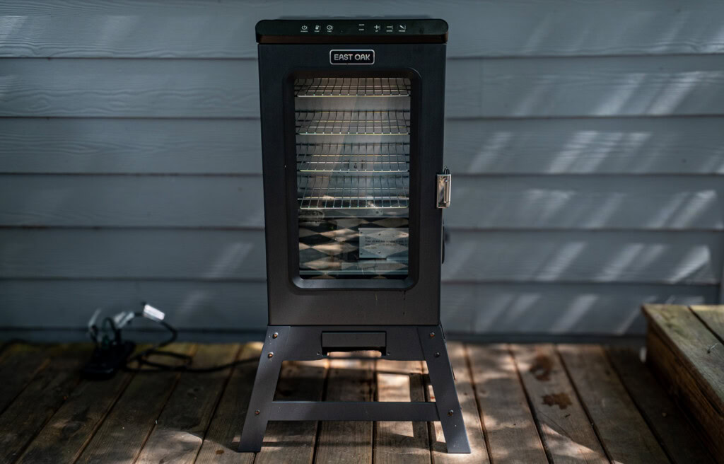 Front view of the East Oak 30-inch electric smoker showing its vertical black metal body, glass viewing window, multiple internal cooking racks, and control panel on top, placed on a wooden deck against a grey siding wall.