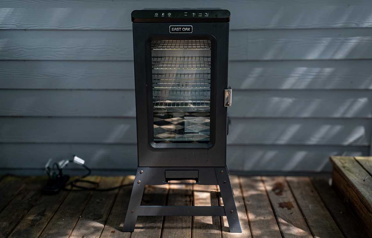 Front view of the East Oak 30-inch electric smoker showing its vertical black metal body, glass viewing window, multiple internal cooking racks, and control panel on top, placed on a wooden deck against a grey siding wall.