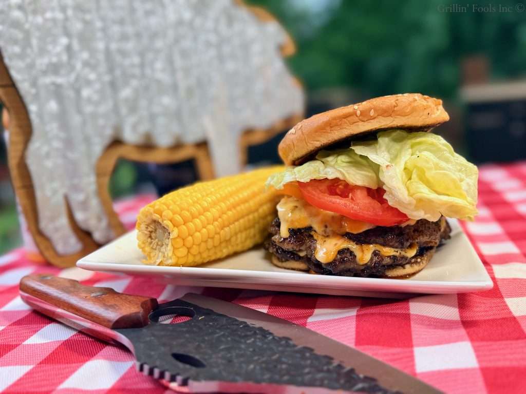 A loaded ground pork smash burger topped with cheese, lettuce, red and yellow tomato on a toasted sesame bun in front of an ear of corn and a metal pig sign. A knife is in the foreground