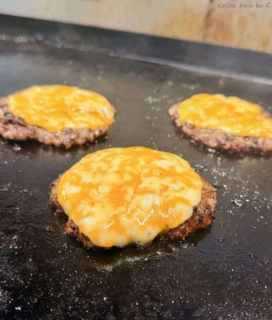 Three cheese topped ground pork smash burger patties on a flat top griddle