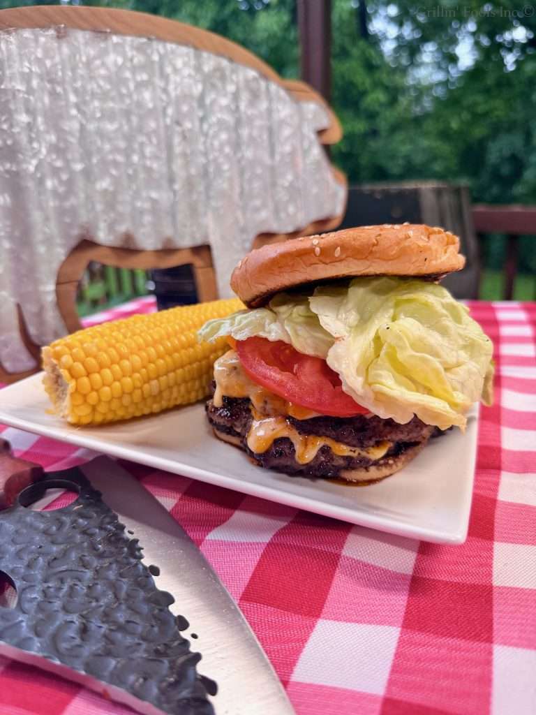 Triple decker, ground pork smash burgers, topped with lettuce and tomato, on a plate with an ear of yellow corn in front of a metal pig sign