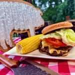 Cheeseburger with lettuce, tomato, and melted cheese on a bun, plated with corn on the cob outdoors on a red checkered tablecloth.