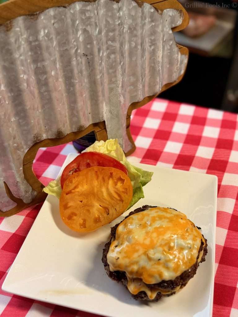 Cheeseburger on a white square plate with melted cheese, tomato slice, and lettuce on a red-and-white checkered tablecloth.