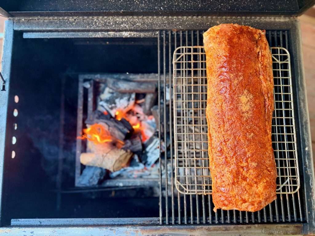 Seasoned pork loin on the upper rack of a grill to the right of a charcoal fire.